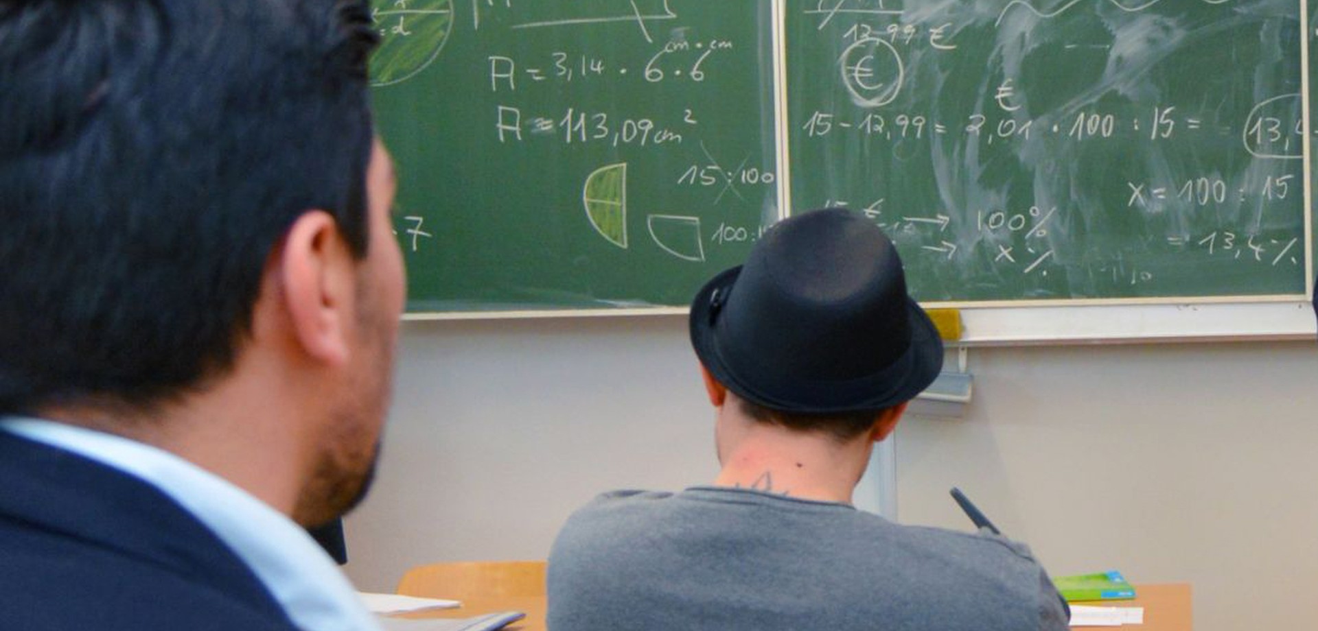 Zwei Männer sitzen an Tischen vor einer Tafel, die mit Formeln beschrieben ist.