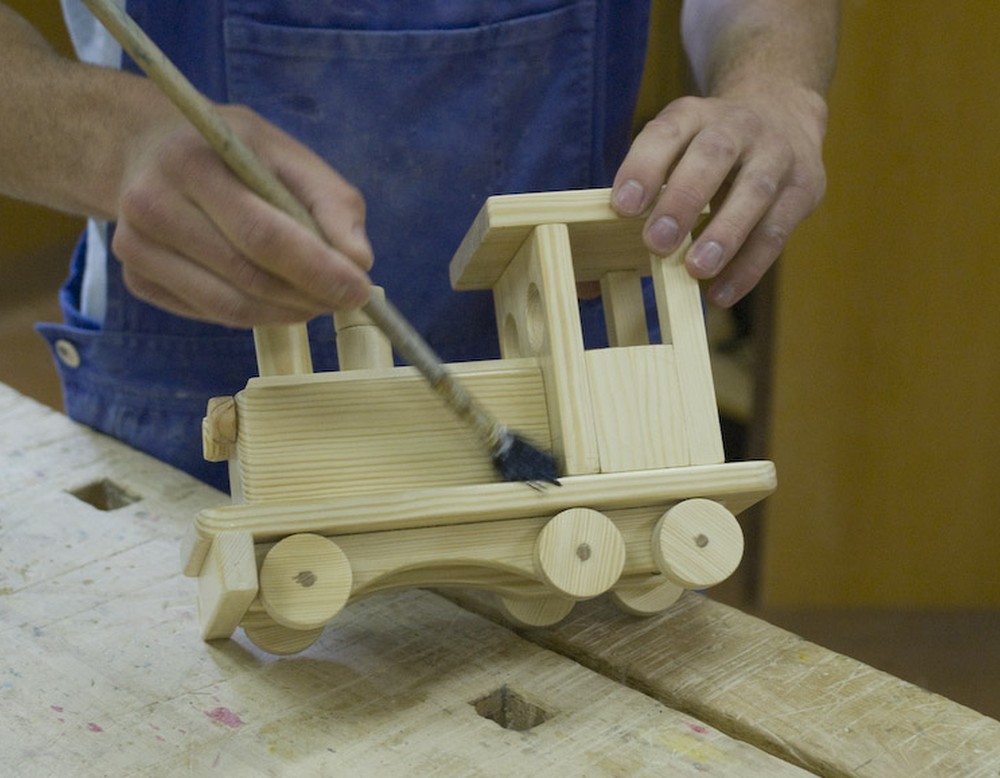 Ein Mann im Blaumann lasiert mit einem Pinsel eine Spielzeuglokomotive aus Holz.