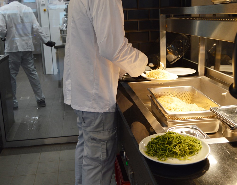 Ein Mann mit Kochjacke steht in der Küche und portoniert Spaghetti mit grüner Soße auf weiße Teller.
